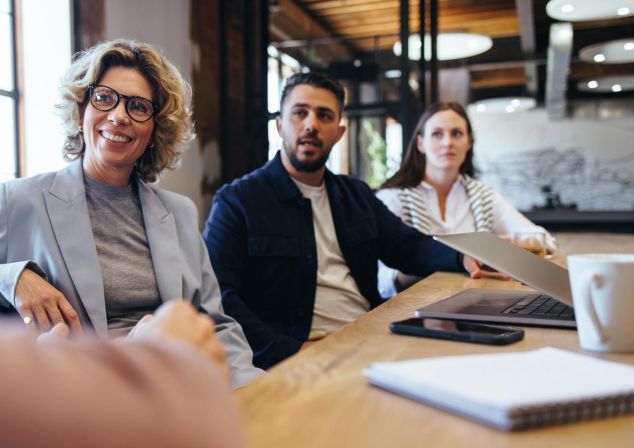 Junge Frau sitzt mit Kollegen am Tisch und schaut fröhlich über den Tisch.