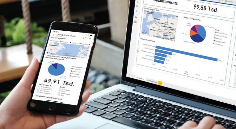 Eine Person sitzt vor einem Laptop, schaut sich ein Power BI Dashboard auf dem Monitor an und vergleicht es mit den Ergebnissen auf einem Handy.