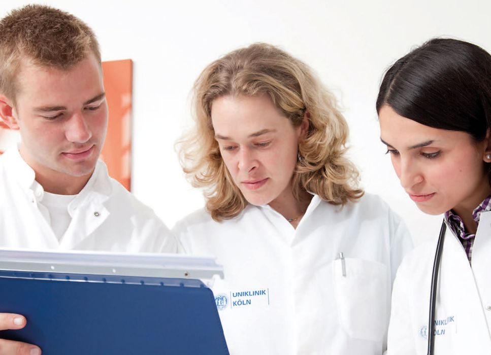 Ärzteteam: Von links ein Mann, und zwei Frauen in einem weißen Kittel mit dem Blick nach unten auf einen Ordner.