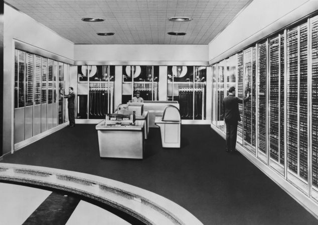 Eine schwarz-weiß Aufnahme aus den 60er Jahren, eine Frau und zwei Männer arbeiten an einem Supercomputer.