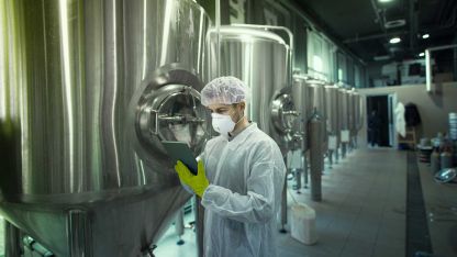 A man with protective equipment and a tablet in his hand stands in front of large stainless steel food containers