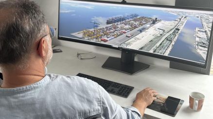 Ein Mann sitzt an einem Schreibtisch und arbeitet vor einem breiten, gebogenen Monitor, auf dem eine Luftaufnahme eines geschäftigen Seehafens mit Containern und Kais zu sehen ist.