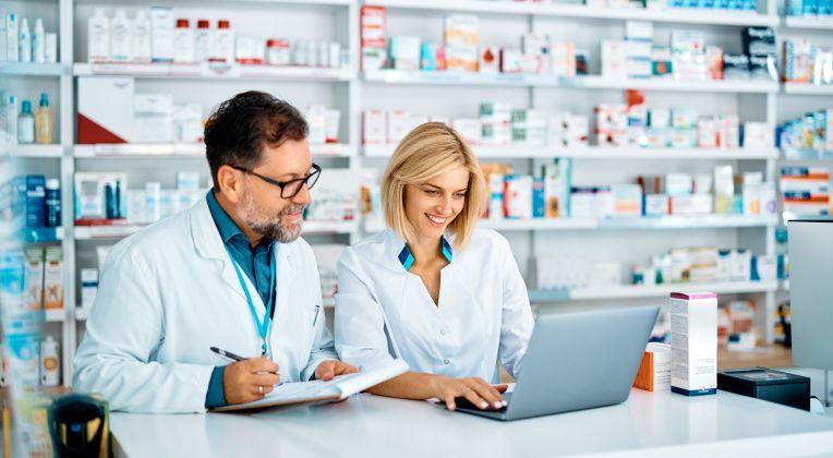 Zwei Apotheker: innen sitzen an einem Tisch in einer Apotheke und gucken auf einen Laptop