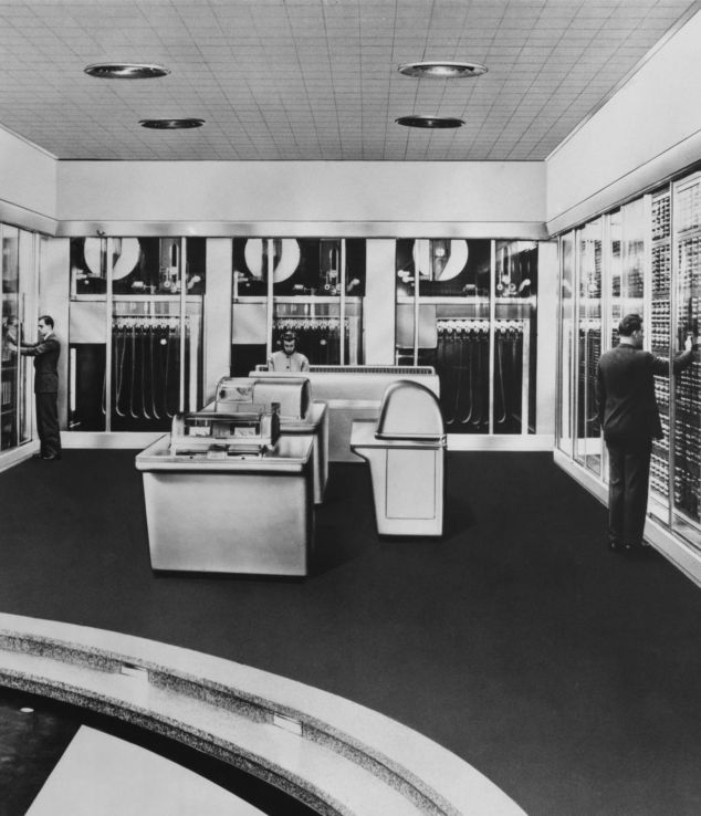 Eine schwarz-weiß Aufnahme aus den 60er Jahren, eine Frau und zwei Männer arbeiten an einem Supercomputer.