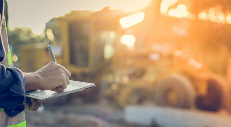 Mann mit Weste und Tablet schreibt Notizen auf einer Baustelle mit LKW im Hintergrund.