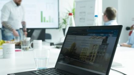 A training participant works on his laptop while our speaker Marcus Wuest gives a presentation with diagrams on a large screen in the background. The training takes place in a modern, light-flooded meeting room.