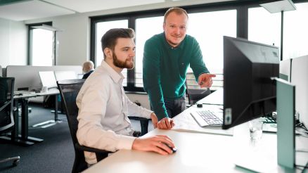 Zwei Mitarbeiter stehen und sitzen an einem Schreibtisch mit Monitor im Büro