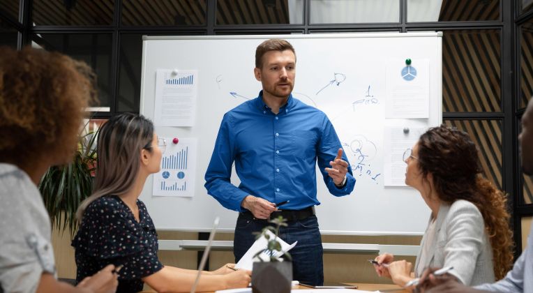 Ein Mann hält an einem Whiteboard eine Präsentation vor einem Team an Kolleg*innen.