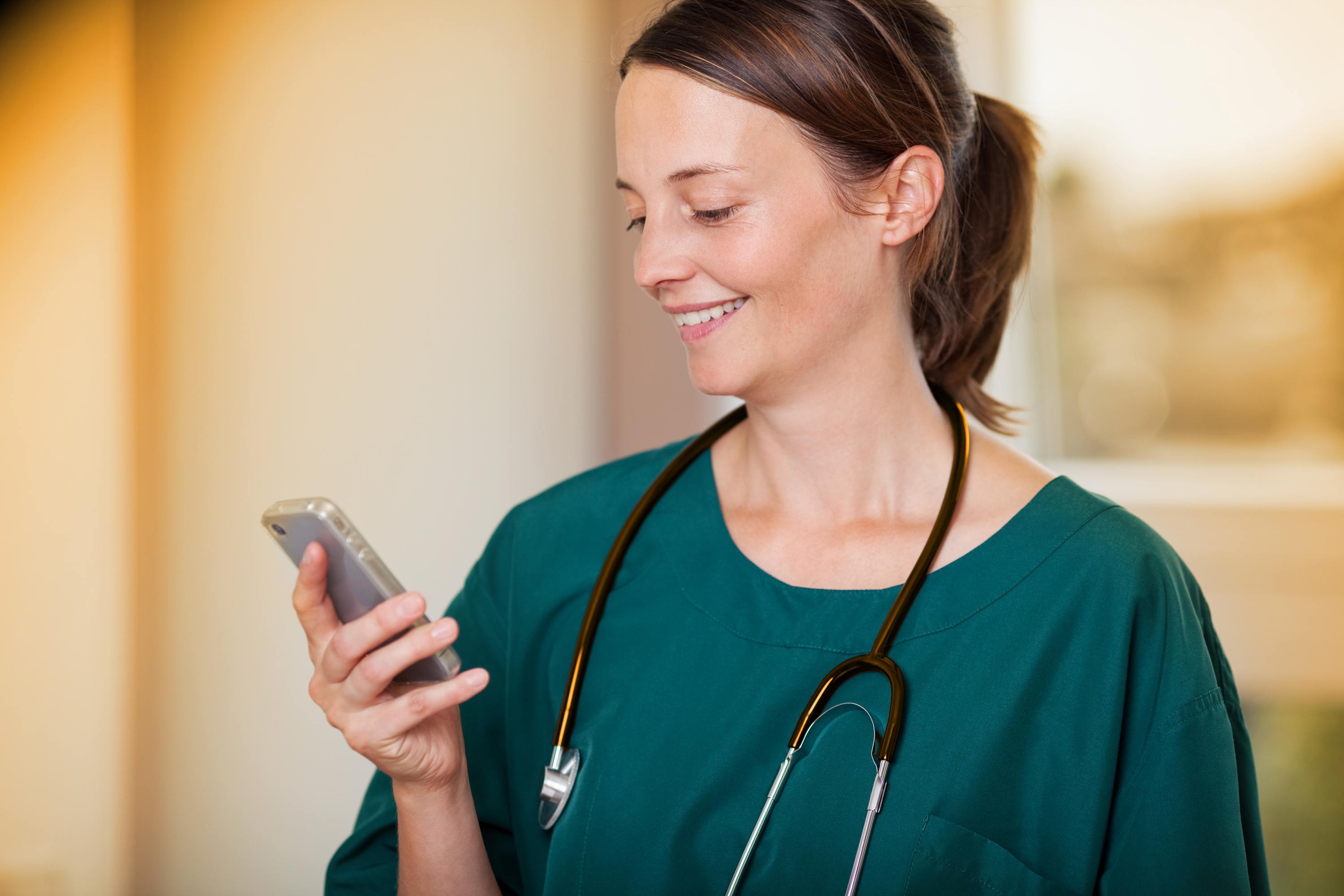 Ärztin mit Stethoskop um den Hals liest lächelnd eine Kurznachricht auf ihrem Smartphone