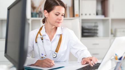 Female doctor in a white coat sitting at a desk and typing on a laptop
