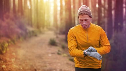 Athlete with orange shirt jogging in the forest takes a look at his heart rate monitor
